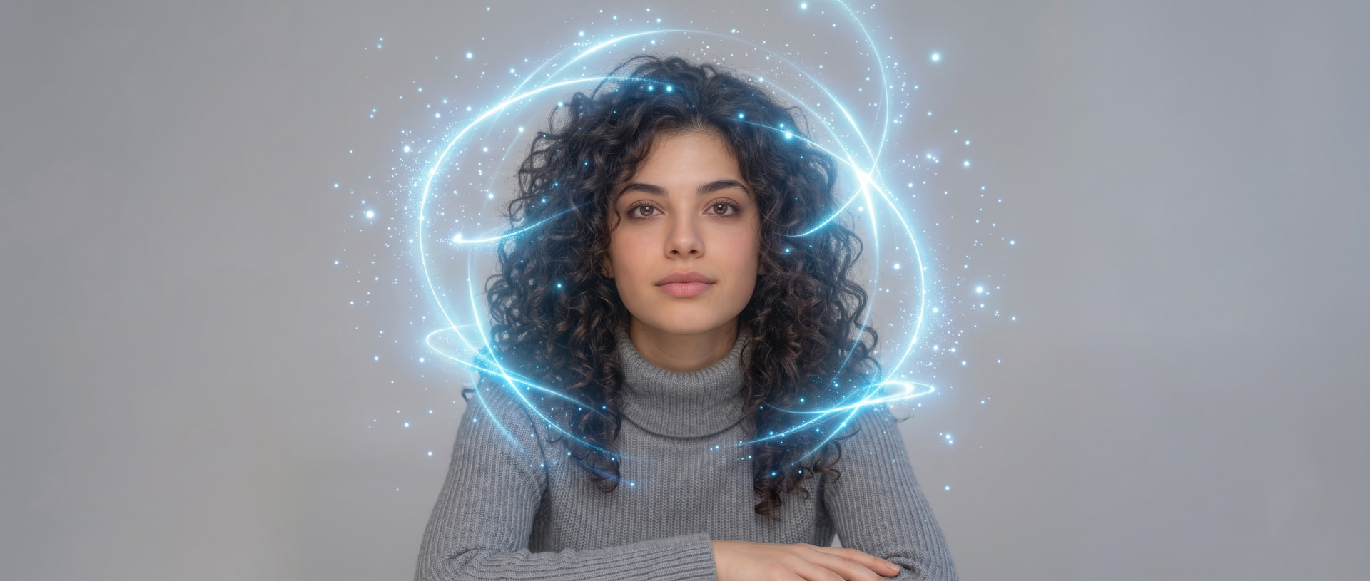 A woman with dark curly hair seated against a soft grey backdrop, luminous blue rings of light spiralling around her head and shoulders